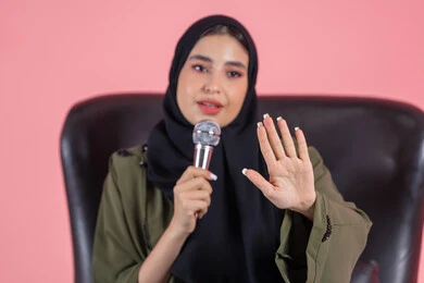 تقديم العروضات المسرحية، الاستمتاع بأداء المواهب الصوتية، مفهوم الفن وتأثيره الايجابي, بورتريه لامرأة اماراتية عربية خليجية محجبة ترتدي عباءة خضراء تجلس على كرسي مريح و تمسك المايكروفون و ترفع يدها بإيماءات الاندماج والتركيز، خلفية وردية