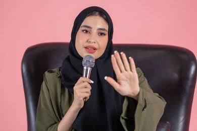 فن الالقاء والخطابة، بدء التدريب الصوتي، تقديم العروض الفنيه و الغنائيه والفقرات الترفيهية، مفهوم الفن وتأثيره الايجابي, بورتريه لامرأة اماراتية عربية خليجية محجبة ترتدي عباءة خضراء تجلس على كرسي مريح و تمسك المايكروفون و ترفع يدها بإيماءات الاندماج والتركيز، خلفية وردية