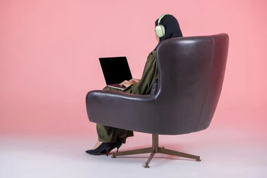 ادارة العمل عن بعد، إنجاز المهام المطلوبة عبر اللابتوب، استخدام جهاز تقني حديث ومتطور, بورتريه من الخلف لامرأة عربية خليجية اماراتية محجبة ترتدي العباءة و سماعات الرأس تجلس على كرسي تعمل على جهاز الحاسوب المحمول، خلفية وردية