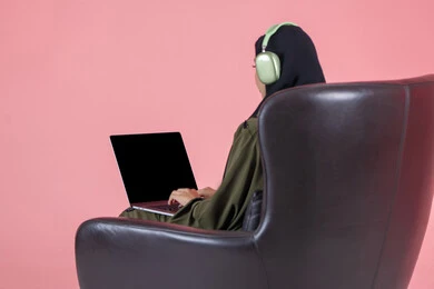 Completing the required tasks via laptop, using a modern and advanced technical device, managing work remotely, a portrait from behind of an Arab Gulf Emirati woman wearing a hijab and an abaya, wearing headphones, sitting on a chair working on a laptop, with a pink background.