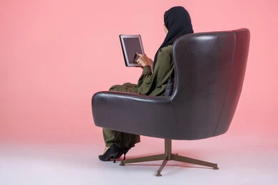 Remote business management, using advanced modern devices and technologies, a portrait from behind of an Arab Gulf Emirati woman wearing a hijab and abaya sitting on a chair working on a tablet, pink background.