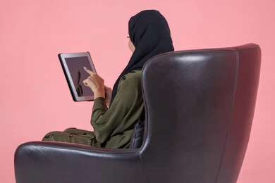 The concept of remote task completion, using advanced modern devices and technologies, a portrait from behind of an Arab Gulf Emirati woman wearing a hijab and abaya sitting on a chair working on a tablet, with a pink background.