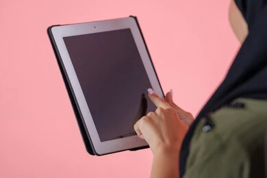 The use of advanced modern technology in professional life through a tablet, browsing social media, a close-up portrait of the hand of an Emirati Arab Gulf woman wearing a hijab working on a tablet, with a pink background.