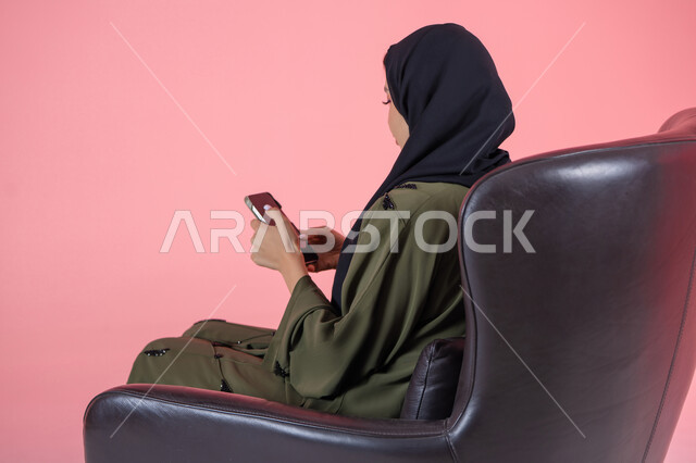 مشاهدة البرامج المفضلة, الاستفادة من خدمات وتطبيقات الموبايل، استخدام الأجهزة والوسائل التقنية الحديثة, بورتريه لامرأة اماراتية عربية خليجية محجبة ترتدي عباءة خضراء تجلس على كرسي مريح و تتصفح وسائل التواصل الاجتماعي عبر الهاتف المحمول, خلفية وردية