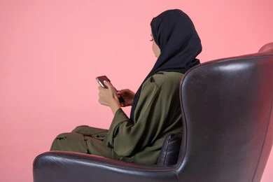 Watching favorite programs, taking advantage of mobile services and applications, using modern technical devices and means, portrait of a veiled Emirati Arab Gulf woman wearing a green abaya sitting on a comfortable chair and browsing social media via mobile phone, pink background