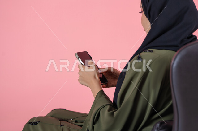 Benefiting from mobile services and applications, using modern technical devices and means, portrait of a veiled Emirati Arab Gulf woman wearing a green abaya sitting on a comfortable chair and browsing social media via mobile phone, pink background
