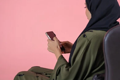 الاستفادة من خدمات وتطبيقات الموبايل، استخدام الأجهزة والوسائل التقنية الحديثة, بورتريه لامرأة اماراتية عربية خليجية محجبة ترتدي عباءة خضراء تجلس على كرسي مريح و تتصفح وسائل التواصل الاجتماعي عبر الهاتف المحمول, خلفية وردية