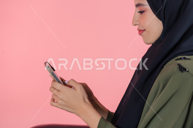 برامج وتطبيقات الجوال المتنوعة، إيماءات السعادة و السرور, استخدام الأجهزة و الوسائل التقنية الحديثة, بورتريه لامرأة اماراتية عربية خليجية محجبة مبتسمة ترتدي عباءة خضراء تجلس على كرسي مريح و تتصفح وسائل التواصل الاجتماعي عبر الهاتف المحمول, خلفية وردية