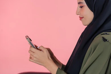 Various mobile programs and applications, gestures of happiness and pleasure, use of modern technical devices and means, portrait of a smiling veiled Emirati Arab Gulf woman wearing a green abaya sitting on a comfortable chair and browsing social media via mobile phone, pink background
