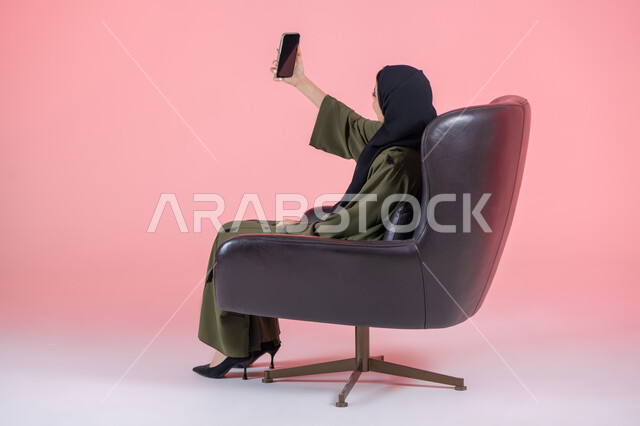 مفهوم التصوير والشهرة عبر برامج التواصل الاجتماعي، استخدام الأجهزة والوسائل التقنية الحديثة, بورتريه لامرأة اماراتية عربية خليجية محجبة ترتدي عباءة خضراء تجلس على كرسي مريح تقوم بالتقاط صورة سيلفي عبر الهاتف المحمول، خلفية وردية