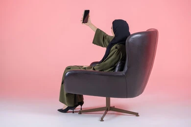مفهوم التصوير والشهرة عبر برامج التواصل الاجتماعي، استخدام الأجهزة والوسائل التقنية الحديثة, بورتريه لامرأة اماراتية عربية خليجية محجبة ترتدي عباءة خضراء تجلس على كرسي مريح تقوم بالتقاط صورة سيلفي عبر الهاتف المحمول، خلفية وردية
