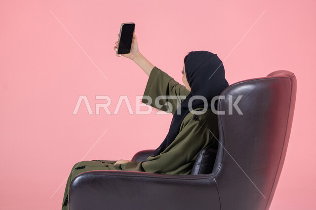 التقاط الصور التذكارية، مشاركة اللحظات السعيدة على مواقع التواصل الاجتماعي، استخدام الأجهزة والوسائل التقنية الحديثة, بورتريه لامرأة اماراتية عربية خليجية محجبة ترتدي عباءة خضراء تجلس على كرسي مريح تقوم بالتقاط صورة سيلفي عبر الهاتف المحمول، خلفية وردية