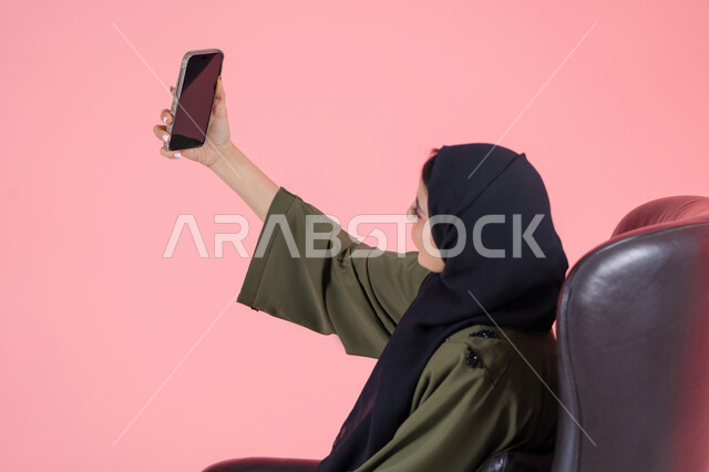 مشاركة اللحظات السعيدة على مواقع التواصل الاجتماعي، استخدام الأجهزة والوسائل التقنية الحديثة, بورتريه لامرأة اماراتية عربية خليجية محجبة ترتدي عباءة خضراء تجلس على كرسي مريح تقوم بالتقاط صورة سيلفي تذكارية عبر الهاتف المحمول، خلفية وردية
