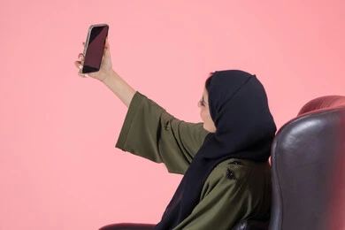 Sharing happy moments on social media, using modern technology, portrait of a veiled Emirati Gulf Arab woman wearing a green abaya sitting on a comfortable chair taking a souvenir selfie with a mobile phone, pink background
