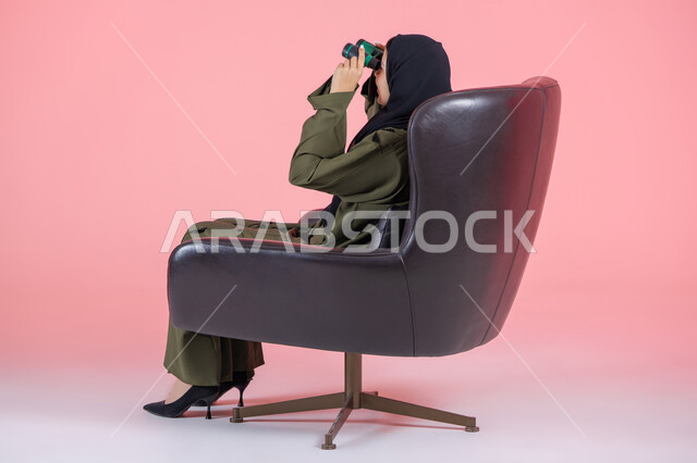 البحث عن شيء ما باستخدام الناظور، مفهوم رؤية الأشياء البعيدة، التخطيط و وضع الأهداف المستقبلية، بورتريه من الجانب لامرأة اماراتية عربية خليجية محجبة ترتدي عباءة خضراء و تجلس على كرسي مريح و تنظر من خلال التلسكوب, خلفية وردية