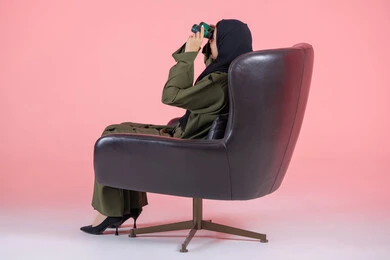 البحث عن شيء ما باستخدام الناظور، مفهوم رؤية الأشياء البعيدة، التخطيط و وضع الأهداف المستقبلية، بورتريه من الجانب لامرأة اماراتية عربية خليجية محجبة ترتدي عباءة خضراء و تجلس على كرسي مريح و تنظر من خلال التلسكوب, خلفية وردية
