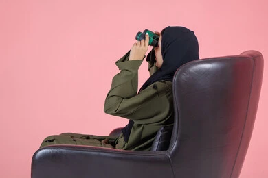 استخدام المنظار ثنائي العينين لمشاهدة الأشياء البعيدة، التخطيط و وضع الأهداف المستقبلية، بورتريه من الجانب لامرأة اماراتية عربية خليجية محجبة ترتدي عباءة خضراء و تجلس على كرسي مريح و تنظر من خلال التلسكوب, خلفية وردية