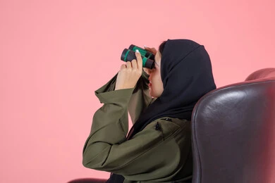 مفهوم تقريب الأشياء, استخدام المنظار ثنائي العينين لمشاهدة الأشياء البعيدة، التخطيط و وضع الأهداف المستقبلية، بورتريه من الجانب لامرأة اماراتية عربية خليجية محجبة ترتدي عباءة خضراء و تجلس على كرسي مريح و تنظر من خلال التلسكوب, خلفية وردية