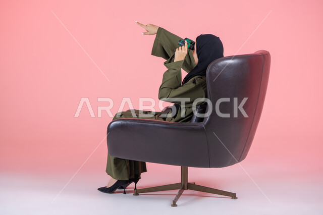 Seeing distant objects through a telescope, future planning for goals and ambitions, pointing with the index finger at something, side portrait of a veiled Emirati Arab Gulf woman wearing a green abaya and sitting on a comfortable chair and looking through a viewfinder, pink background
