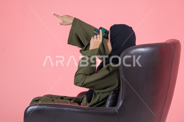 حب المغامرة و الاستكشاف، رؤية الاشياء البعيدة عبر التلسكوب، التخطيط المستقبلي للأهداف و الطموحات، الإشارة بإصبع السبابة الى شيء ما, بورتريه من الجانب لامرأة اماراتية عربية خليجية محجبة ترتدي عباءة خضراء و تجلس على كرسي مريح و تنظر من خلال منظار الرؤية, خلفية وردية