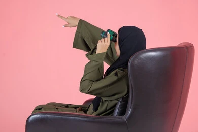 حب المغامرة و الاستكشاف، رؤية الاشياء البعيدة عبر التلسكوب، التخطيط المستقبلي للأهداف و الطموحات، الإشارة بإصبع السبابة الى شيء ما, بورتريه من الجانب لامرأة اماراتية عربية خليجية محجبة ترتدي عباءة خضراء و تجلس على كرسي مريح و تنظر من خلال منظار الرؤية, خلفية وردية