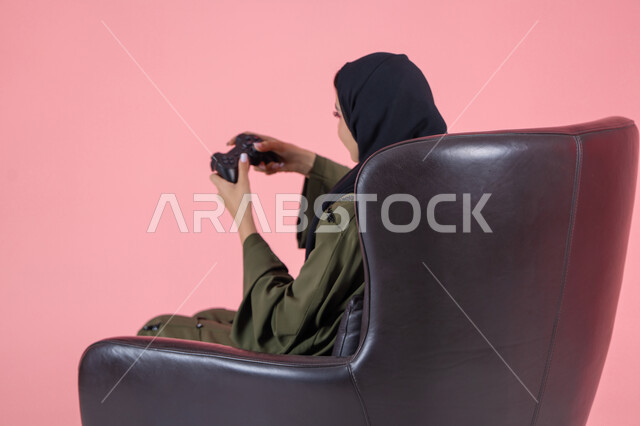 أنشطة ترفيهية ممتعة, قضاء أوقات الفراغ بلعب البلايستيشن, بورتريه من الجانب لامرأة اماراتية عربية خليجية محجبة ترتدي عباءة خضراء و تجلس على كرسي مريح وتحمل بيدها عصا التحكم و تلعب بألعاب الفيديو, خلفية وردية