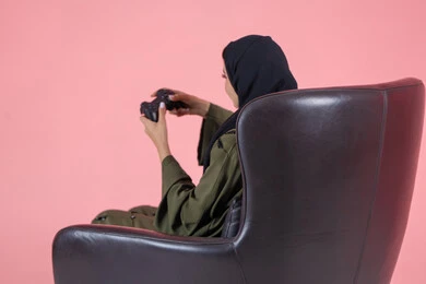 أنشطة ترفيهية ممتعة, قضاء أوقات الفراغ بلعب البلايستيشن, بورتريه من الجانب لامرأة اماراتية عربية خليجية محجبة ترتدي عباءة خضراء و تجلس على كرسي مريح وتحمل بيدها عصا التحكم و تلعب بألعاب الفيديو, خلفية وردية