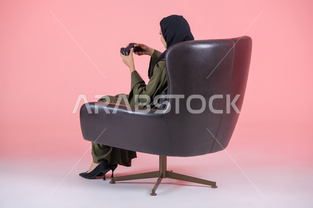 تقنيات حديثة مسلية, أنشطة ترفيهية ممتعة, قضاء أوقات الفراغ بلعب البلايستيشن, بورتريه من الجانب لامرأة اماراتية عربية خليجية محجبة ترتدي عباءة خضراء و تجلس على كرسي مريح وتحمل بيدها عصا التحكم و تلعب بألعاب الفيديو, خلفية وردية