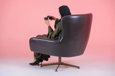 تقنيات حديثة مسلية, أنشطة ترفيهية ممتعة, قضاء أوقات الفراغ بلعب البلايستيشن, بورتريه من الجانب لامرأة اماراتية عربية خليجية محجبة ترتدي عباءة خضراء و تجلس على كرسي مريح وتحمل بيدها عصا التحكم و تلعب بألعاب الفيديو, خلفية وردية