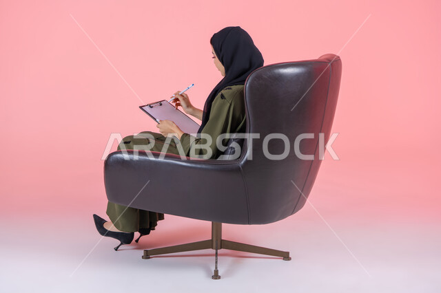 كتابة التقارير و الابحاث على الورق, تدوين وتسجيل المعلومات, تنظيم وإدارة الأعمال, كتابة المهام والأهداف اليومية, بورتريه من الجانب لامرأة اماراتية عربية خليجية محجبة ترتدي عباءة خضراء تجلس على كرسي مريح وتحمل بيدها أوراق وملفات خاصة بالعمل, خلفية وردية