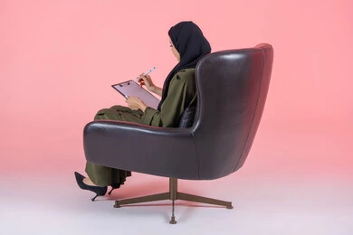 كتابة التقارير و الابحاث على الورق, تدوين وتسجيل المعلومات, تنظيم وإدارة الأعمال, كتابة المهام والأهداف اليومية, بورتريه من الجانب لامرأة اماراتية عربية خليجية محجبة ترتدي عباءة خضراء تجلس على كرسي مريح وتحمل بيدها أوراق وملفات خاصة بالعمل, خلفية وردية