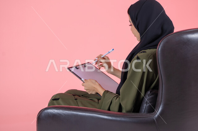 تدوين وتسجيل المعلومات على الورق, تنظيم وإدارة الأعمال, كتابة المهام والأهداف اليومية, بورتريه من الخلف لامرأة اماراتية عربية خليجية محجبة ترتدي عباءة خضراء تجلس على كرسي مريح وتحمل بيدها أوراق وملفات خاصة بالعمل, خلفية وردية