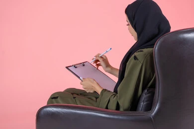 تدوين وتسجيل المعلومات على الورق, تنظيم وإدارة الأعمال, كتابة المهام والأهداف اليومية, بورتريه من الخلف لامرأة اماراتية عربية خليجية محجبة ترتدي عباءة خضراء تجلس على كرسي مريح وتحمل بيدها أوراق وملفات خاصة بالعمل, خلفية وردية