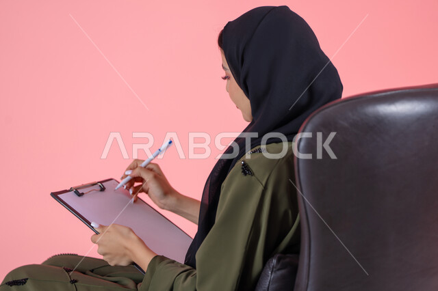 كتابة المهام والأهداف اليومية,  تدوين وتسجيل المعلومات على الورق, تنظيم وإدارة الأعمال, بورتريه من الخلف لامرأة اماراتية عربية خليجية محجبة ترتدي عباءة خضراء تجلس على كرسي مريح وتحمل بيدها أوراق وملفات خاصة بالعمل, خلفية وردية