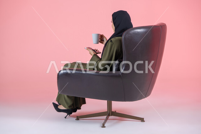 Enjoying delicious healthy drinks, side portrait of a veiled Emirati Arab Gulf woman wearing a green abaya and sitting on a comfortable chair holding a mobile phone and a cup of warm drink, communicating with family and friends, pink background
