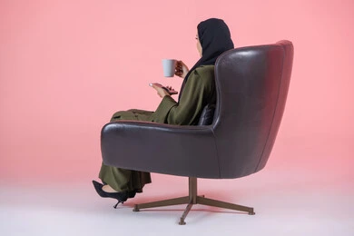 Enjoying delicious healthy drinks, side portrait of a veiled Emirati Arab Gulf woman wearing a green abaya and sitting on a comfortable chair holding a mobile phone and a cup of warm drink, communicating with family and friends, pink background