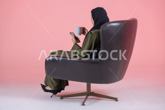 تناول المشروب الساخن والأعشاب الشتوية الدافئة, قضاء الوقت باحتساء المشروبات المفضلة, بورتريه من الجانب لامرأة اماراتية عربية خليجية محجبة ترتدي عباءة خضراء و تجلس على كرسي مريح تمسك بيدها الهاتف المحمول والكوب, خلفية وردية