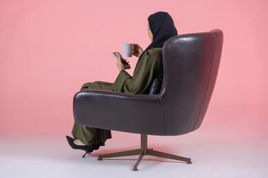 Drinking a hot drink and warm winter herbs, spending time sipping your favorite drinks, side portrait of a veiled Emirati Arab Gulf woman wearing a green abaya and sitting on a comfortable chair holding a mobile phone and a cup, pink background