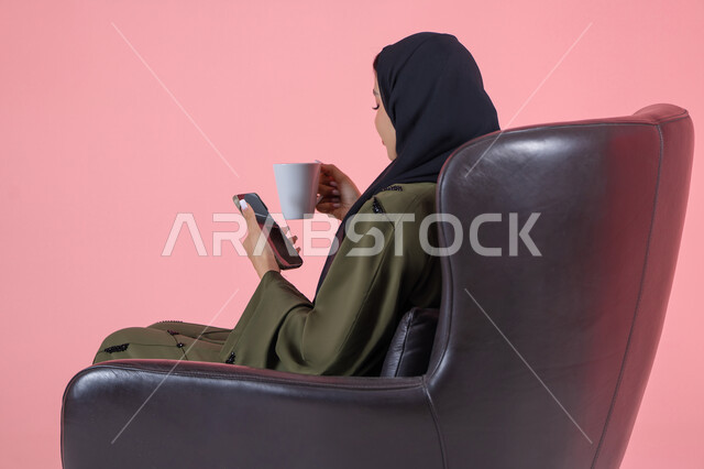 قضاء الوقت باحتساء المشروبات المفضلة, بورتريه من الجانب لامرأة اماراتية عربية خليجية محجبة ترتدي عباءة خضراء و تجلس على كرسي مريح تمسك بيدها الهاتف المحمول و كوب من المشروب الدافئ,  التواصل مع الأهل والأصدقاء، خلفية وردية