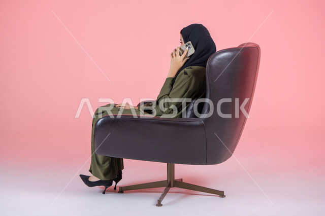 التواصل والاطمئنان على الأصدقاء, إجراء اتصال هاتفي عبر الجوال, استخدام جهاز تقني حديث و متطور, بورتريه من الجانب لامرأة اماراتية عربية خليجية محجبة ترتدي عباءة خضراء و تجلس على كرسي مريح و تستخدم الهاتف في التواصل مع الأقارب, مشاركة الأخبار والأحاديث, خلفية وردية