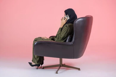 Communicating and checking up on friends, making a phone call via mobile, using a modern and advanced technical device, a side portrait of a veiled Emirati Arab Gulf woman wearing a green abaya and sitting on a comfortable chair and using the phone to communicate with relatives, sharing news and conversations, pink background