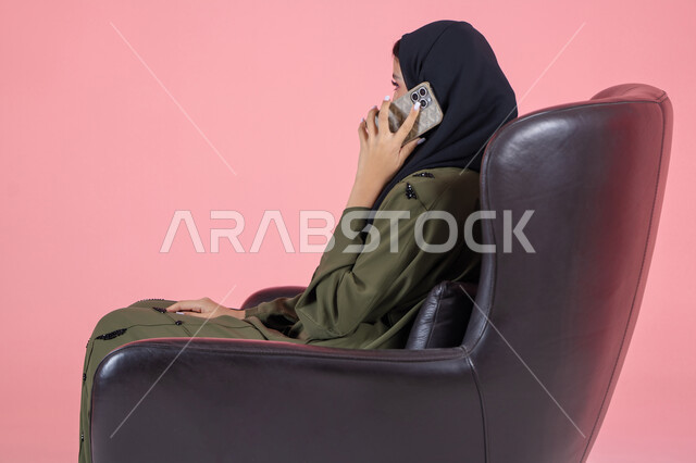 دردشات مع الاصدقاء، إجراء اتصال هاتفي عبر الجوال, استخدام جهاز تقني حديث و متطور, بورتريه من الجانب لامرأة اماراتية عربية خليجية محجبة ترتدي عباءة خضراء و تجلس على كرسي مريح تستخدم الهاتف في التواصل مع الأقارب, مشاركة الأخبار والأحاديث, خلفية وردية