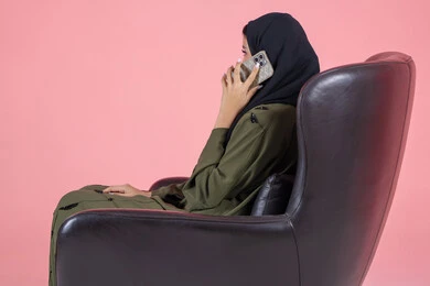Chatting with friends, making a phone call via mobile, using a modern and advanced technical device, a side portrait of a veiled Emirati Arab Gulf woman wearing a green abaya and sitting on a comfortable chair using the phone to communicate with relatives, sharing news and conversations, pink background