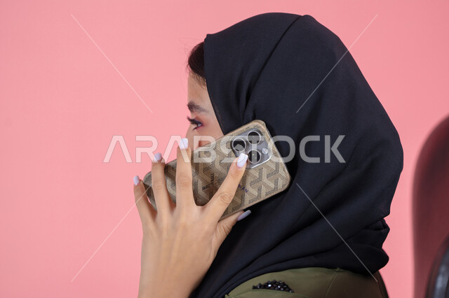 إجراء اتصال هاتفي عبر الجوال, استخدام جهاز تقني حديث و متطور, محادثات و دردشات مع الاصدقاء، بورتريه من الجانب لامرأة اماراتية عربية خليجية محجبة ترتدي عباءة خضراء و تستخدم الهاتف في التواصل مع الأقارب, خلفية وردية