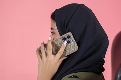 إجراء اتصال هاتفي عبر الجوال, استخدام جهاز تقني حديث و متطور, محادثات و دردشات مع الاصدقاء، بورتريه من الجانب لامرأة اماراتية عربية خليجية محجبة ترتدي عباءة خضراء و تستخدم الهاتف في التواصل مع الأقارب, خلفية وردية