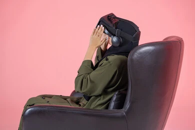 Entertainment by watching through virtual reality glasses, spending leisure time in an enjoyable way, interacting and simulating the imaginary virtual world, enjoying and spending recreational time playing video games, modern technologies and technological devices, a side portrait of a veiled Emirati Arab Gulf woman wearing a green abaya and putting on VR glasses, pink background