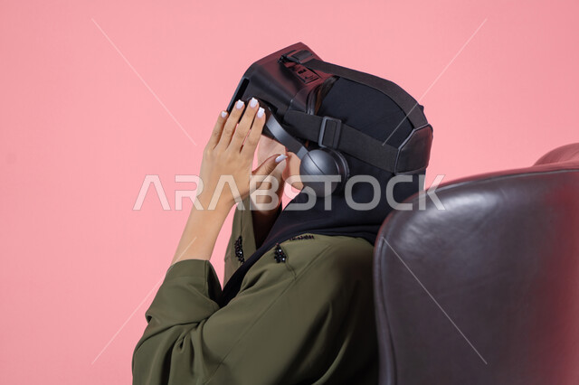 Spending leisure time in a fun way, interacting and simulating the imaginary virtual world, enjoying and spending recreational time playing video games, modern technologies and technological devices, close-up portrait of a veiled Emirati Arab Gulf woman wearing a green abaya and VR glasses, pink background