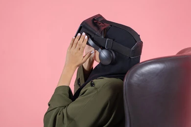 Spending leisure time in a fun way, interacting and simulating the imaginary virtual world, enjoying and spending recreational time playing video games, modern technologies and technological devices, close-up portrait of a veiled Emirati Arab Gulf woman wearing a green abaya and VR glasses, pink background