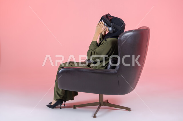 Merging entertainment with the imaginary world, technical and technological development, enjoyment and viewing through virtual reality glasses, a side portrait of a veiled Emirati Arab Gulf woman wearing a green abaya, putting on VR glasses and sitting on a comfortable chair, pink background