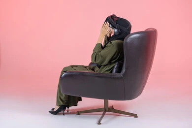 Merging entertainment with the imaginary world, technical and technological development, enjoyment and viewing through virtual reality glasses, a side portrait of a veiled Emirati Arab Gulf woman wearing a green abaya, putting on VR glasses and sitting on a comfortable chair, pink background